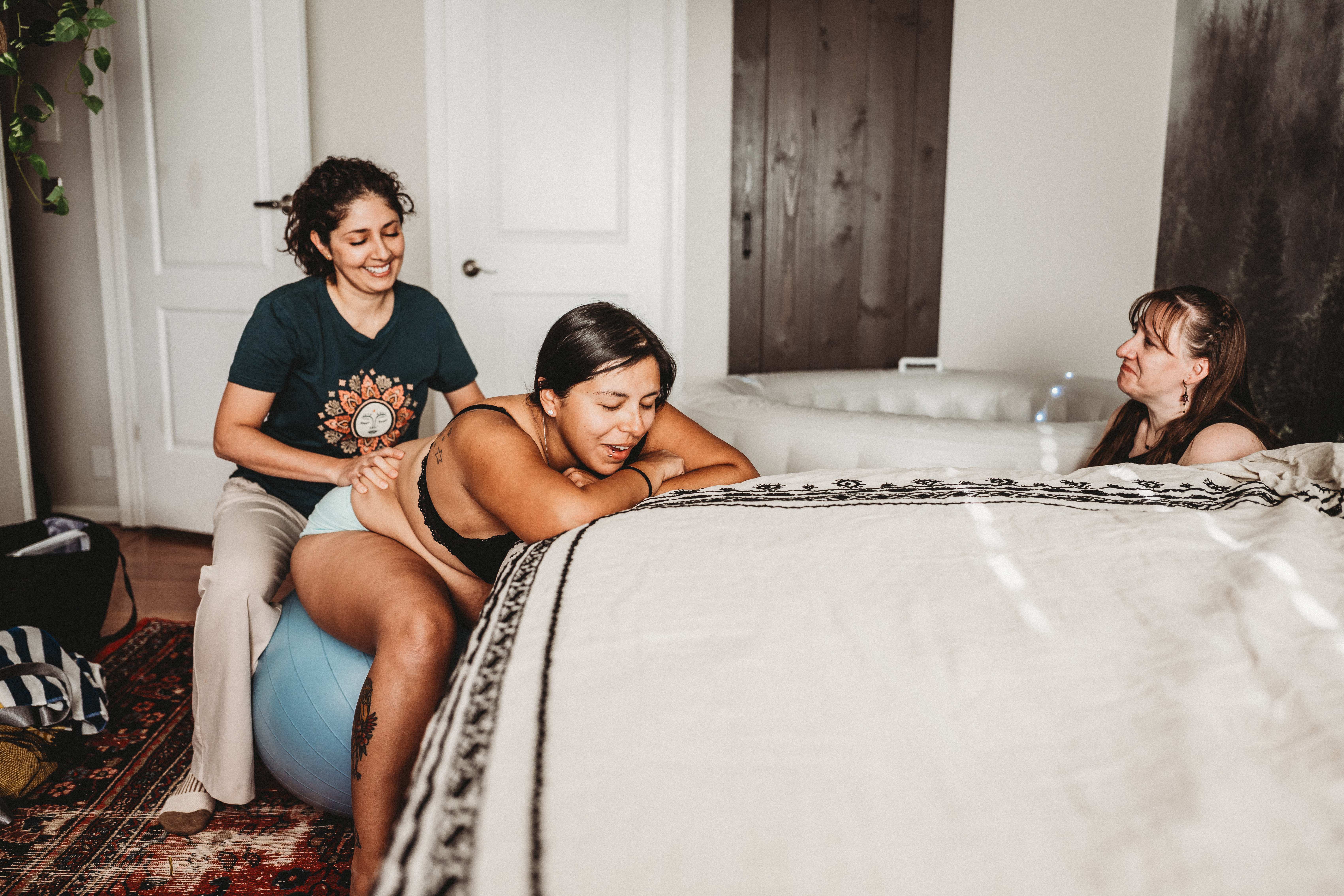 Maribel supporting a laboring mother leaning over the bed as the midwife looks on